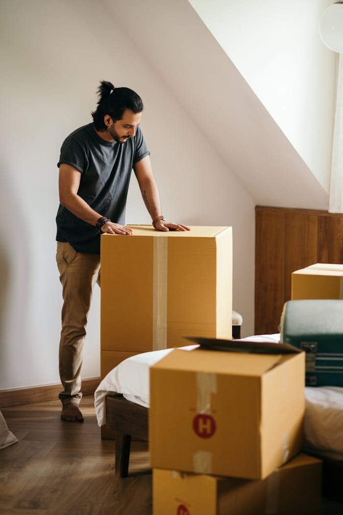 about-img Ethnic man packing belongings in cardboard boxes while standing in modern apartment with white wall and looking down while preparing for relocation